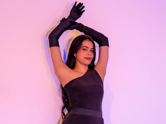 Alana Blake mit langen, dunklen Haaren, trägt ein schwarzes Kleid mit einem Schulterträger und schwarze Handschuhe, die Arme sind erhoben, die Beleuchtung ist lila und rosa, sie lächelt, zeigt ihre Brüste leicht und wirkt sexy..