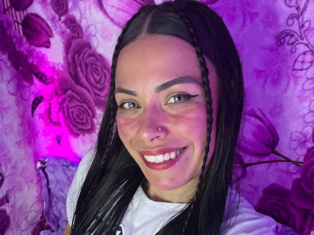 ZoeLux has long black braids, a nose ring, and a big smile with red lipstick. She's wearing a white shirt, and her tits are small. She's cute with purple lighting.