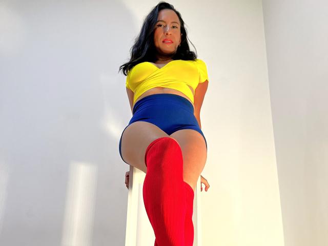 Jade Foster, con i capelli neri, una maglietta gialla, pantaloncini blu e calze rosse, è in piedi con una gamba sollevata, mostrando il seno e i glutei, e guarda verso il basso la fotocamera..