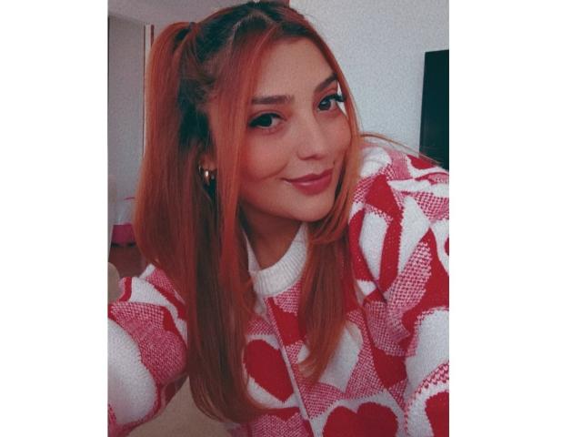BiancaRey, con cabello rojo-anaranjado en coletas, vistiendo un suéter rosa y blanco con estampado de corazones, sonriendo a la cámara, mostrando su linda cara y sus pequeños senos..
