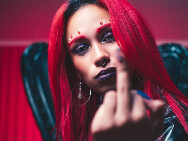 LilithDeveraux, con cabello rojo, maquillaje oscuro y sombra de ojos roja, señalando a la cámara, fondo negro en forma de corazón, aro en la nariz, lápiz labial morado y aretes de plata. Los senos no son visibles..