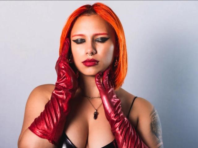 LilithDeveraux mit leuchtend roten Haaren, die glänzende rote Handschuhe trägt, berührt ihr Gesicht, trägt schwarzen Schminke, einen Nasenring, einen tätowierten Arm, hat einen Ausschnitt in einem schwarzen Top, trägt roten Lippenstift und blickt nach unten..