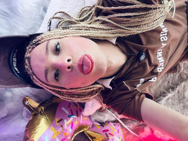 AaronFinn with blonde braids, pouty red lips, brown shirt, and baseball cap, lying on a fluffy white surface, next to a gold and pink donut-shaped pillow.