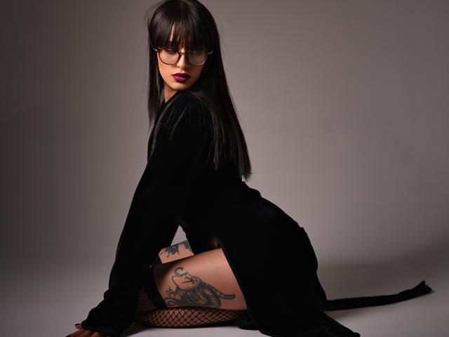 TatsumiLee, with black dress, glasses, and long straight hair, sits on floor, showing tattooed thigh and fishnet stockings, with pink lipstick and serious expression, in dark setting.