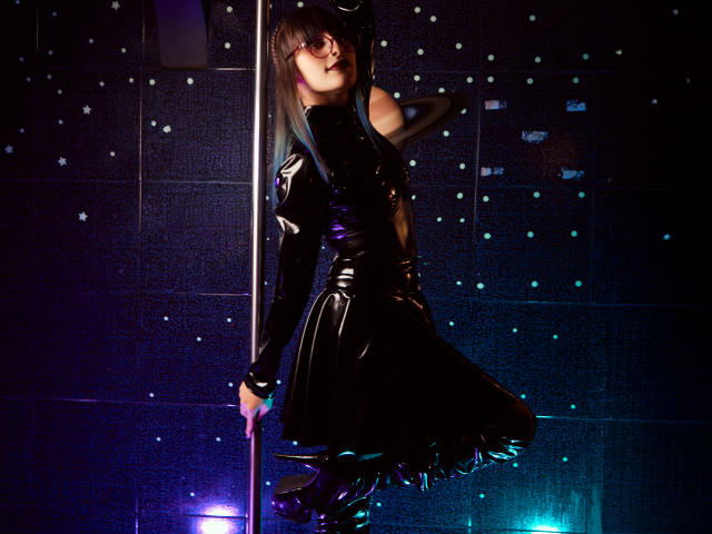 TatsumiLee in a shiny black latex dress, standing by a pole, holding it with one hand, looking back with a sultry expression, blue and purple lights on.