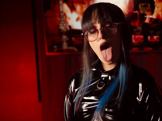 TatsumiLee mit schwarzem Latextop, blauen Haarspitzen und Brille, Zunge heraus, roter Hintergrund, trägt eine Brille mit rotem Rahmen, sieht sexy und verspielt aus..