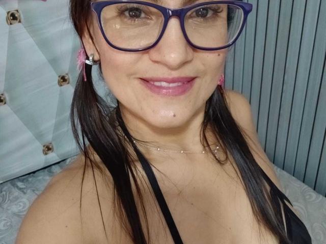 SolaraDubois with dark pigtails, purple glasses, pink lipstick, black top, and a small mole under her right eye, smiling at the camera. She's wearing pink earrings and a necklace.