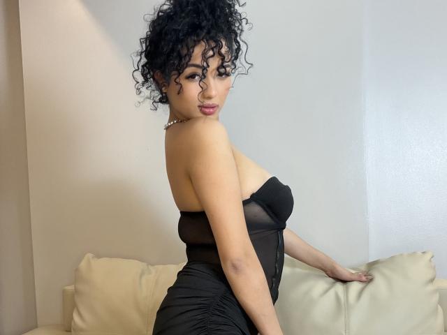 AuroraFerretti with curly black hair, wearing a black strapless dress, showing her tits, sitting on a beige couch, looking over her shoulder, pearl necklace.