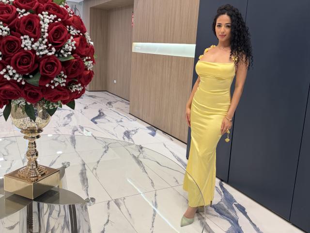 Aurora Ferretti, avec ses cheveux noirs bouclés, portant une robe jaune, se tient à côté d'une table en verre sur laquelle se trouve un bouquet de roses rouges, des fleurs blanches et un vase en or..