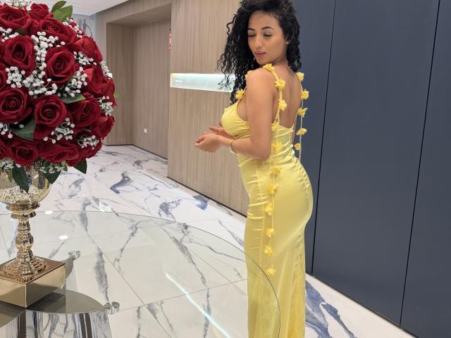 AuroraFerretti in a yellow dress with yellow flower accents, looking over her shoulder, standing near a glass table with a red rose bouquet. Her ass is prominent.