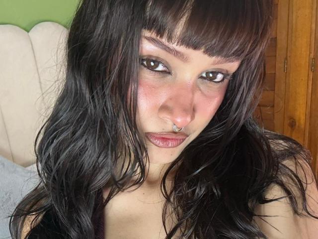 CandyyClair has dark, wavy hair, brown eyes, and a septum piercing. She's looking at the camera with a slight smile, showing her pinkish-red lips. Her skin is fair.