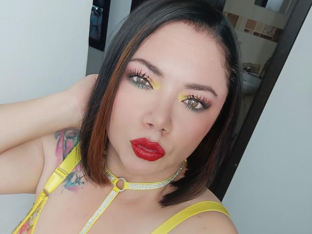 SamanthaVenus with red lipstick, yellow eye makeup, short black-brown hair, yellow choker, and visible tattoo on her right shoulder, looking at the camera with a pouty expression.