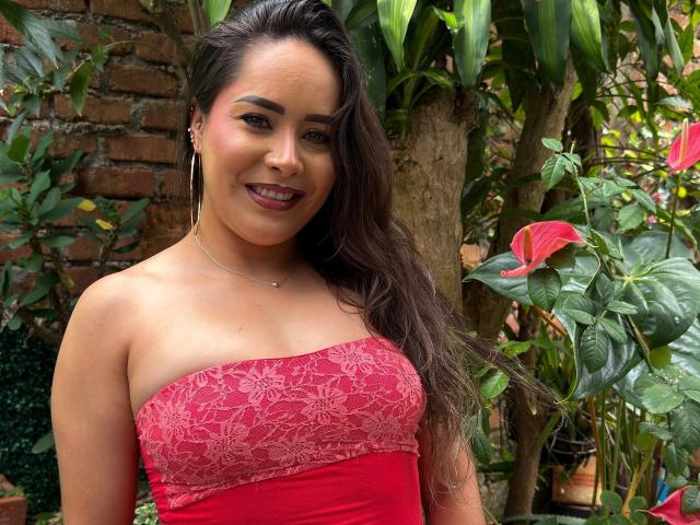 CamilaBardot met lang donker haar, een rode kanten jurk zonder bandjes, lachend, met oorringen, bruine ogen, subtiele make-up, staand in de buurt van groene planten, een rode bloem op de achtergrond, middelgrote borsten, schattig..