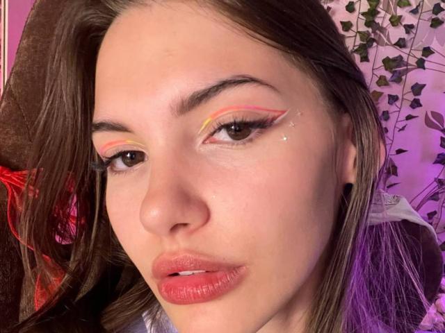 Vonda Bojko, com delineador rosa e amarelo, lábios rosados levemente entreabertos, olhos castanhos e cabelo castanho liso, usando alargadores de orelha pretos, em um ambiente iluminado em tons de rosa..