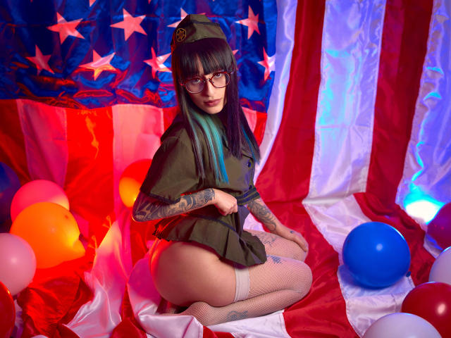 TatsumiLee mit Brille, schwarzem Haar, einem grünen Hut, einem grünen Oberteil, ohne Hose, die ihren Hintern zeigt, mit Tattoos auf den Armen, weißen Fischernetzstrümpfen, kniend auf einer amerikanischen Flagge, umgeben von Ballons..