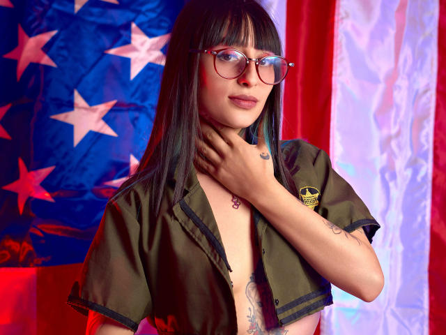 TatsumiLee, avec ses longs cheveux noirs, ses lunettes, et une chemise verte de style militaire ouverte, dévoilant sa poitrine et ses tatouages, tenant son cou devant un fond avec une étoile rouge et bleue..
