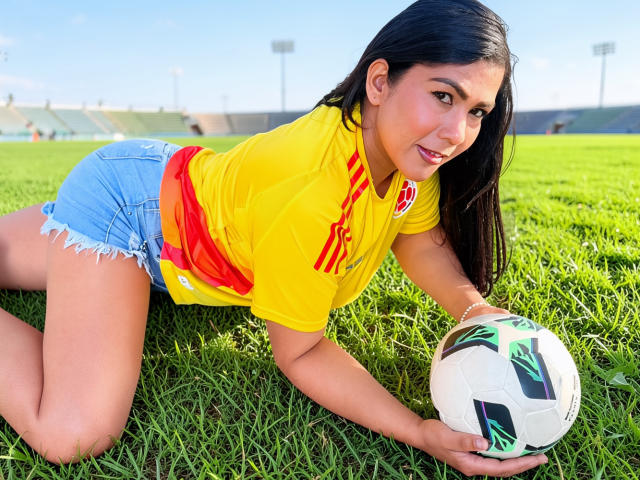IsadoraVex, avec de longs cheveux noirs, un maillot de foot jaune avec des rayures rouges, un short en jean bleu et une ceinture rouge, est allongée sur l'herbe verte, tenant un ballon de foot et souriant à la caméra..