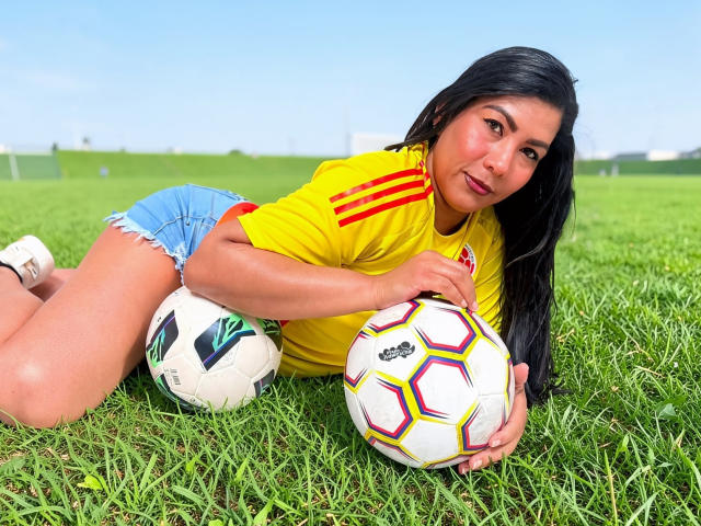 IsadoraVex ligt op het gras, gekleed in een geel voetbalsok, blauwe denim shorts, en houdt twee voetballen vast, waarvan er één wit is met een groen logo, en de andere kleurrijk, met zwart haar en een bruine huid..