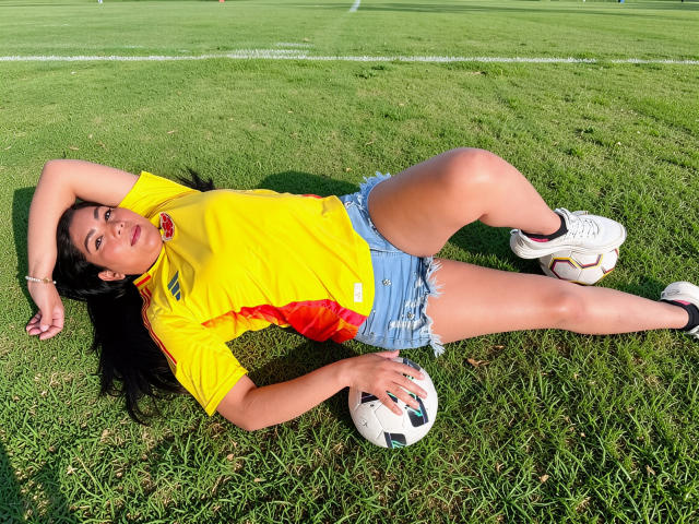 IsadoraVex est allongée sur l'herbe, les bras au-dessus de la tête, portant une chemise jaune, un short bleu, des chaussures de foot blanches, et tenant un ballon de foot, ses fesses légèrement relevées, de longs cheveux noirs éparpillés..