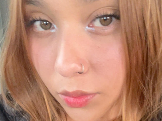 HeraJuno has a cute, serious face with brown eyes, light skin, and pink lips. She has straight, brown hair and a small nose ring. Her lips are slightly pouty.