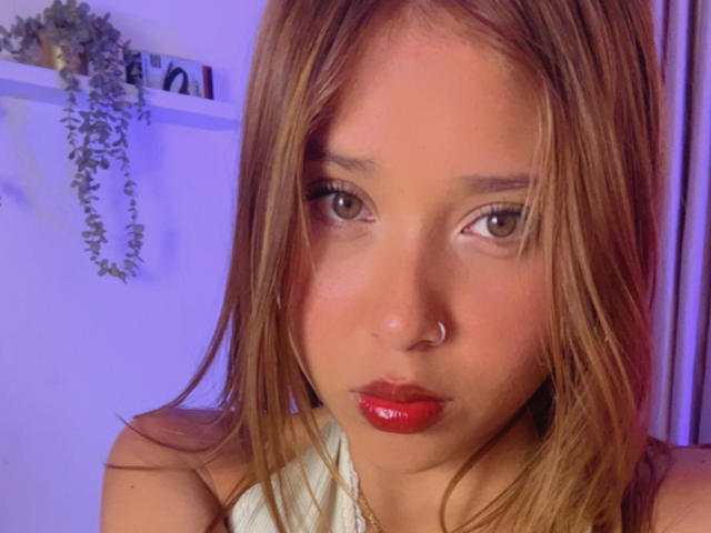 HeraJuno with shiny red lips, nose ring, and brown hair, looking at camera. Her makeup is subtle, and she has a cute, slightly pouty expression. Her tits are small.