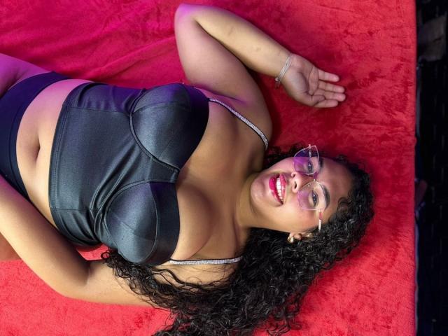AshleyJenner, laying on red fabric, wearing black leather bra, showing big tits, curly black hair, glasses, red lipstick, black panties, right arm raised, left arm resting.