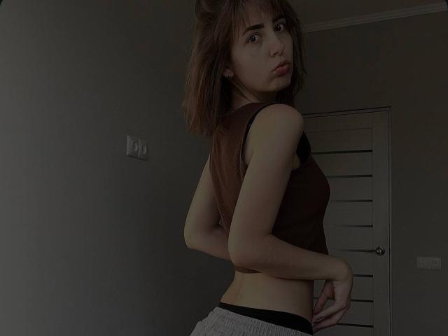 Tora Stambaugh, con un corte de pelo bob castaño, mirando por encima del hombro, vestida con una camiseta marrón sin mangas, pantalones grises, con una mano en la cadera, mostrando su cintura delgada, pechos pequeños y una ligera expresión de puchero..