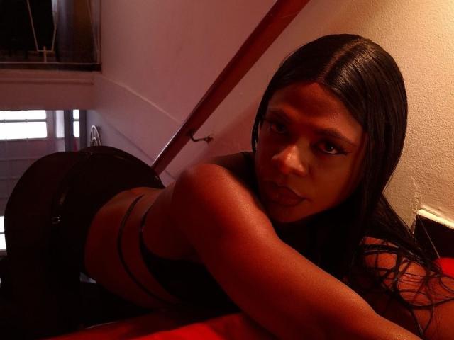 Venus Miller, avec de longs cheveux noirs, regardant l'appareil photo, allongée sur une surface rouge, portant de la lingerie noire, ses fesses relevées, et son visage légèrement incliné, dans un éclairage tamisé..