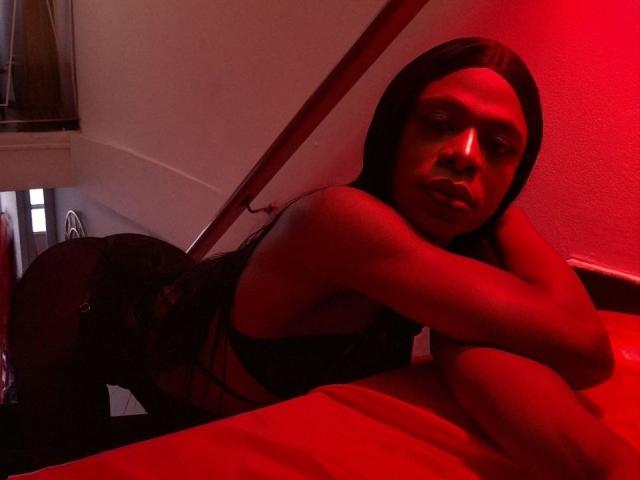 VenusMiller with long black hair, red-lit skin, lying on a bed, wearing a black bra, looking at the camera, her ass slightly raised, arm resting on the bed.