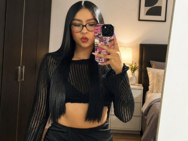 ChloeMoretti with long black hair, glasses, and red lipstick, wearing a sheer black long-sleeve top and black pants, taking a mirror selfie with a pink phone case.