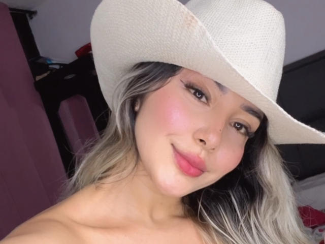 KimmyPinkX con un rubor rosa claro, un sombrero de vaquera blanco, cabello gris y rubio, y lápiz labial rosa, sonriendo a la cámara, mostrando sus hombros desnudos y un anillo nasal adorable..