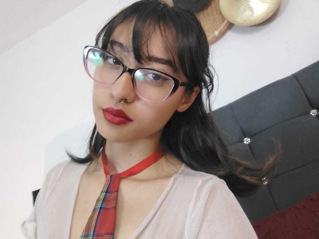CamiVargas with glasses, red nose ring, red lipstick, red tie, plaid, and sheer top, showing her tits, black hair, and red tie. Bedroom setting, looking at camera.