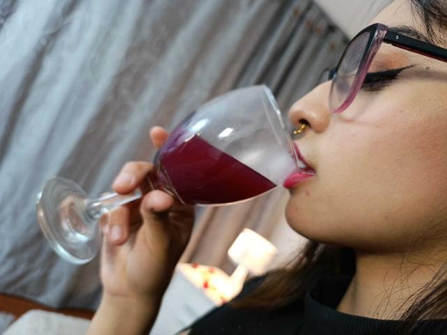 Cami Vargas trinkt Rotwein aus einem großen Glas, trägt eine Brille mit schwarzem Rahmen und ein schwarzes Oberteil, mit pinkem Lippenstift und einem Nasenring. Ihre rechte Hand hält das Glas..