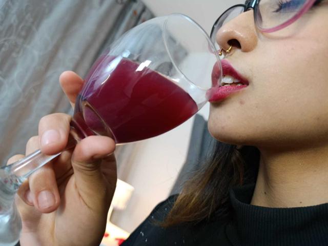 CamiVargas sips red wine from a glass, nose pierced, wearing glasses, black turtleneck, pink lipstick, and brown hair. Her lips are around the glass's rim.