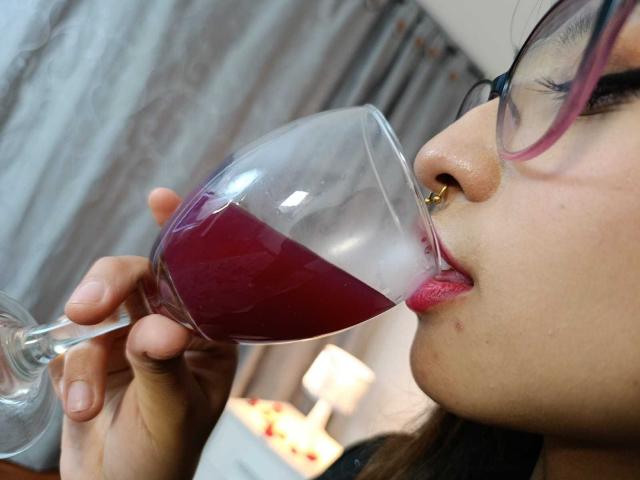 CamiVargas with glasses, nose ring, and pink lipstick, sipping purple wine from a glass, her tongue slightly out, showing off her pink lips and nose ring.