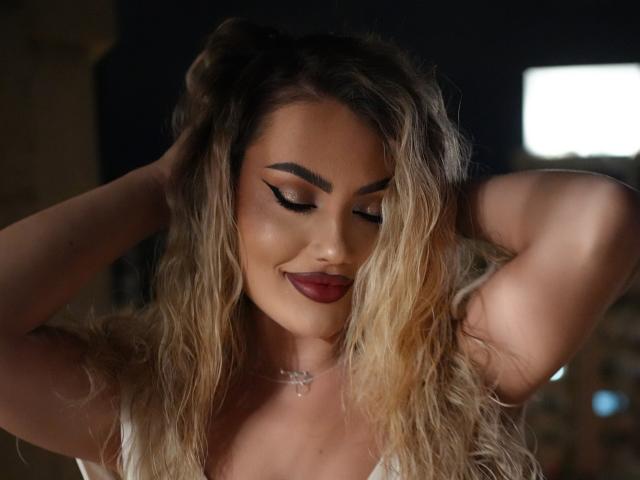 CelineHuddson with wavy blonde hair, dark lipstick, and winged eyeliner, smiling with arms raised, showing her tits in a low-cut top. She's wearing a cross necklace.