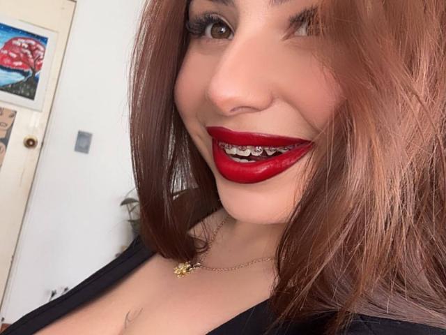 VenusxMusa with red lipstick, showing off her teeth, has brown hair, wearing a black top, and a gold necklace, smiling at the camera. Her tits are partially visible.