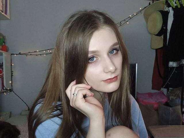 Jaimie Liley, con cabello castaño largo, ojos azules y un piercing en la nariz, vestida con una camisa azul, sosteniendo su barbilla, mirando a la cámara, en una habitación con luz tenue..