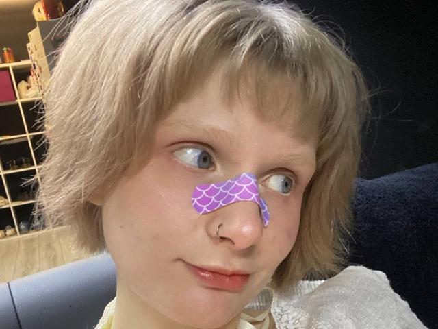 JaimieLiley ha i capelli biondi corti, un naso con un adesivo viola a forma di ragnatela e un piccolo anello al naso. JaimieLiley guarda di lato con un leggero sorriso..