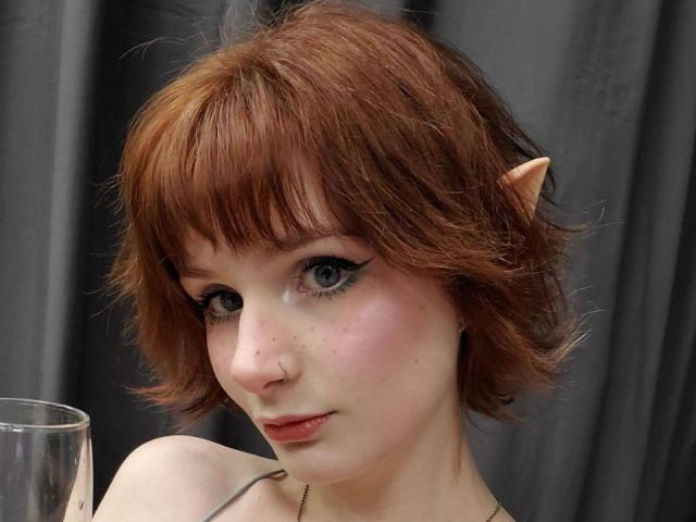 JaimieLiley tem cabelo curto e ruivo, com uma orelha de elfo falsa, olhos azuis, delineador preto e sardas. Ela está segurando um copo, olhando ligeiramente para baixo com um sorriso discreto..