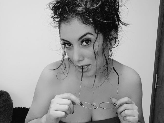 Foto em preto e branco de VickKolles com cabelo despenteado, segurando óculos com os dentes, olhando para a câmera, mostrando seios grandes, nua, com esmalte de unha escuro..