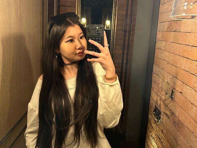 BernadineFray, con lunghi capelli neri, indossa una camicia bianca a maniche lunghe, un choker e orecchie a forma di gatto, e sta scattando un selfie allo specchio con uno smartphone in una stanza poco illuminata..