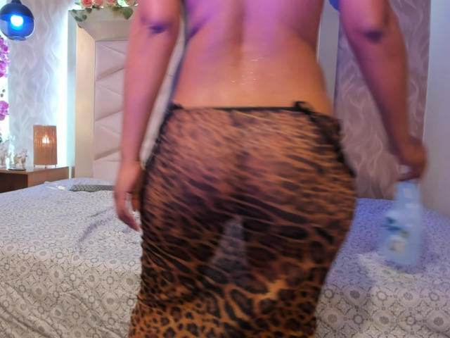 KathaVoxx, sin sujetador, con piel brillante, vistiendo una falda ajustada de estampado de leopardo, sosteniendo una botella de agua, glúteos visibles, tatuaje en el brazo izquierdo, luz azul borrosa en el fondo..