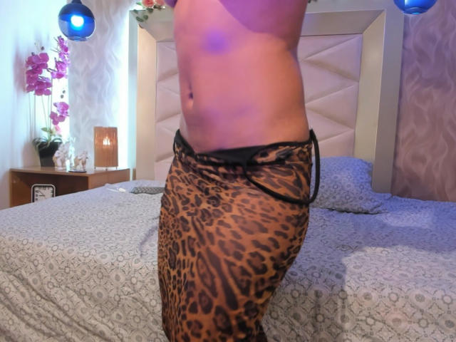 A região do abdômen e as nádegas de KathaVoxx, vestindo calças de estampa de leopardo com detalhes pretos, em um quarto com uma cama de padrão cinza e luzes penduradas azuis..