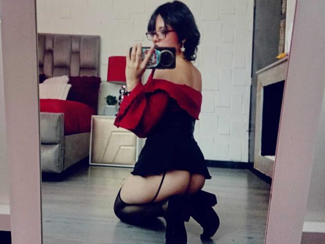 ViolettClay kneeling in front of a mirror, wearing red off-shoulder top, black skirt, garter, and thigh-high stockings, showing her ass, in a bedroom with a bed and lamp.
