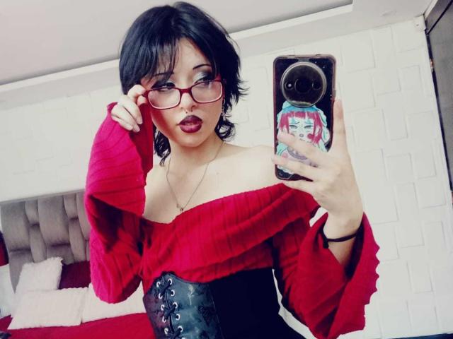 ViolettClay with black hair, glasses, red off-shoulder top, black corset, holding phone with anime sticker, showing nose ring and dark lipstick, in mirror selfie.