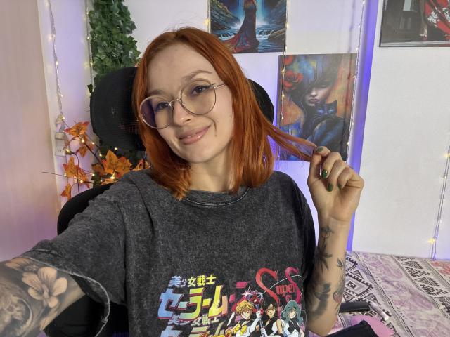 SarahRousen with red hair, glasses, smiling, holding hair, wearing gray graphic tee, tattoos on arms, green nails, sitting in chair, purple and white room, fairy lights.