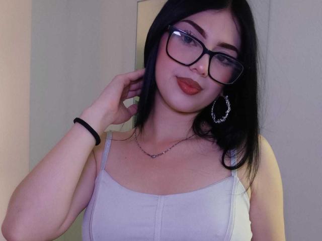 IsabellaaRossi with black glasses, red lipstick, and hoop earrings, wearing a white tank top, showing her tits, hand touching her hair, black bracelet on her wrist.