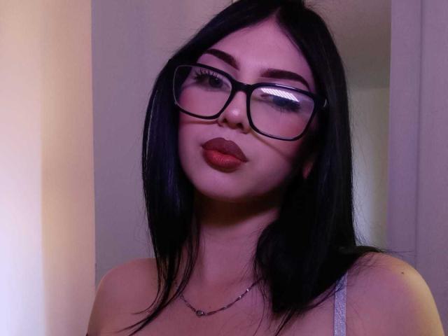IsabellaaRossi with black glasses, red lipstick, and straight black hair, wearing a white strap, looking up with a slight pout, has a silver necklace.