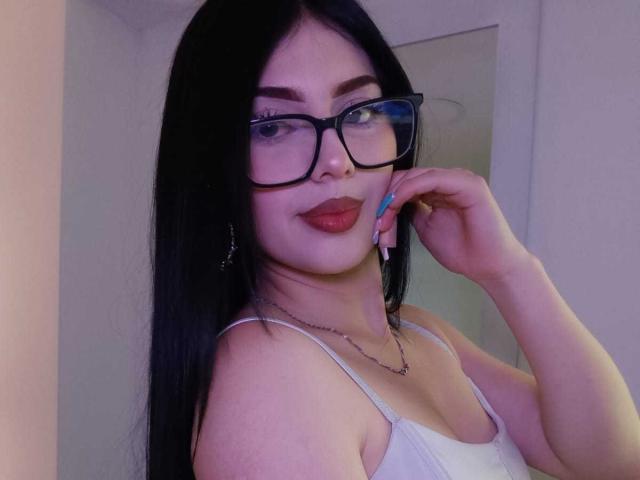 IsabellaaRossi with long black hair, wearing glasses, white tank top, and red lipstick, touching her cheek with blue nails, showing her tits and a small chest tattoo.